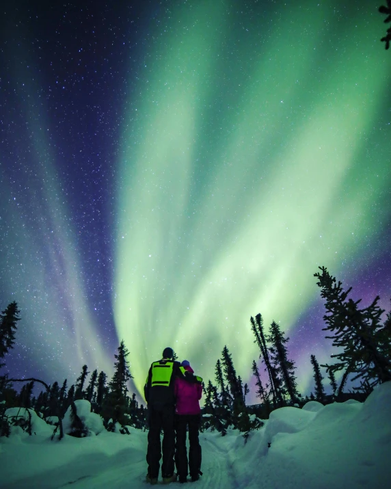 a couple of people that are standing in the snow, pexels contest winner, northern lights, alaska, tall windows lit up, bright vivid colors