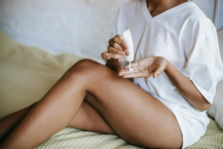 a woman sitting on a bed holding a bottle of lotion, trending on pexels, her tiny hands are on her thighs, contracept, holding an epée, manuka