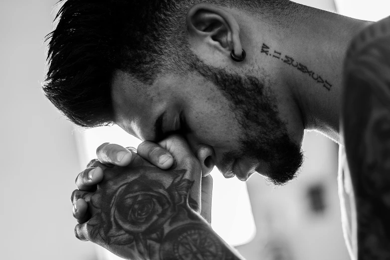 a black and white photo of a man with a tattoo on his arm, a black and white photo, by Matija Jama, pexels, kneeling in prayer, rose tattoo, zayn malik, hands on face