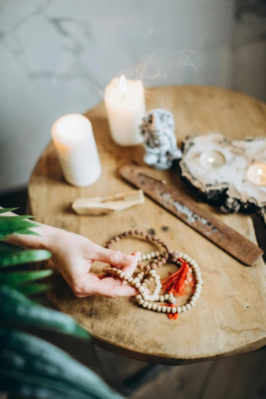 a person holding a necklace on top of a wooden table, a still life, by Julia Pishtar, trending on pexels, white candles, kundalini energy, tassels, spa