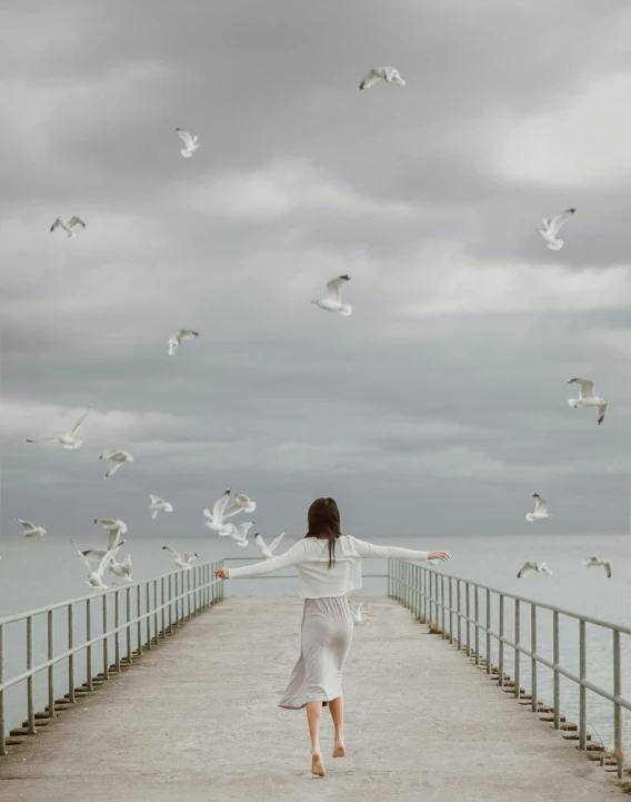a woman standing on a pier surrounded by seagulls, by Lucia Peka, carefree, trending photo, cloudy day, whirling