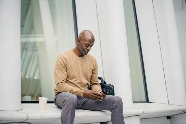 a man sitting on a ledge looking at his cell phone, a photo, he is wearing a brown sweater, lance reddick, digital health, background image