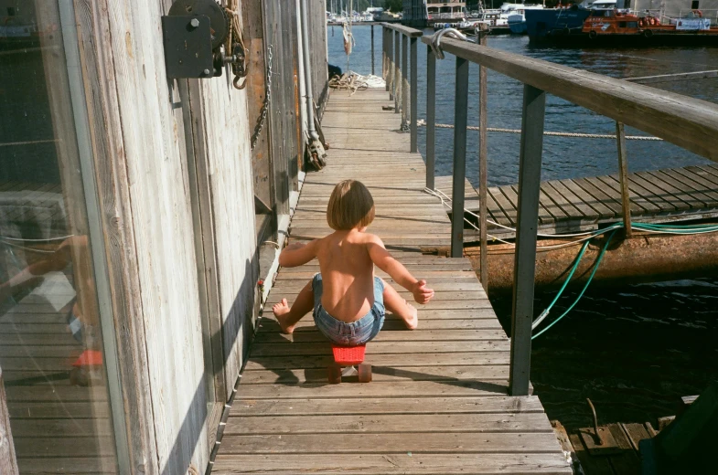 a young boy sitting on a dock next to a body of water, a picture, inspired by Thomas Struth, unsplash, wooden toilets, shipyard, eva elfie, manly