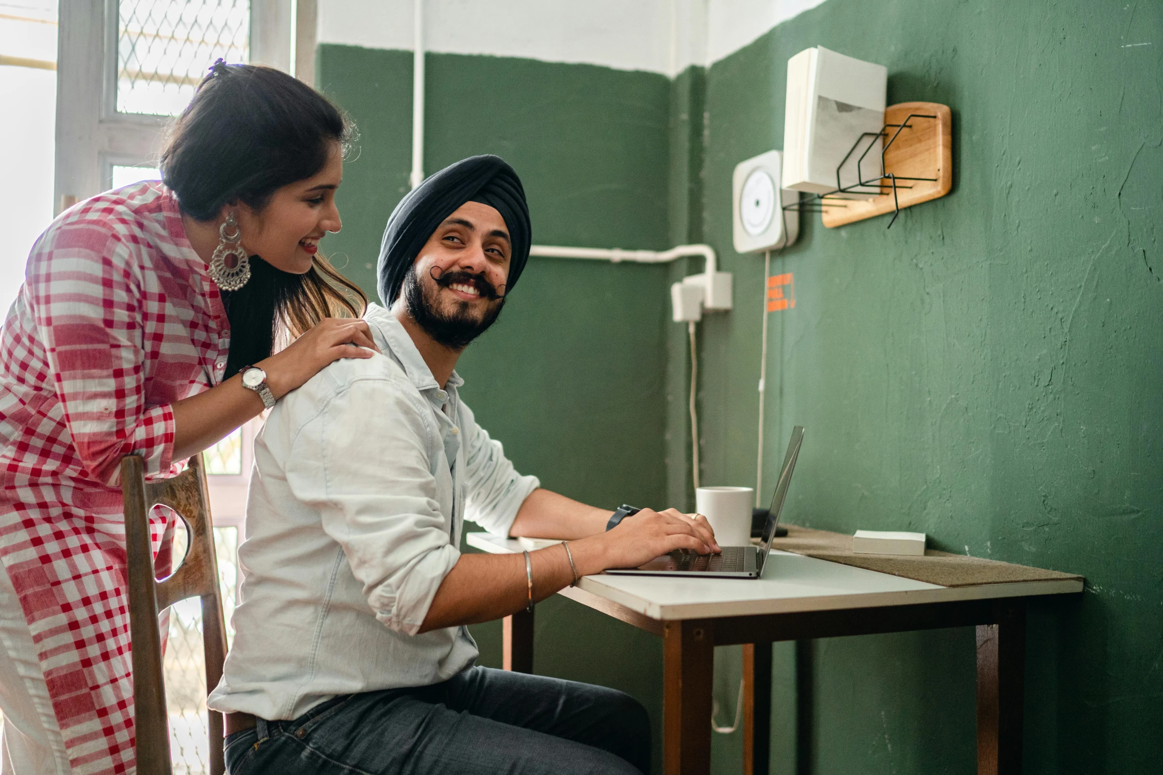 a man and a woman sitting at a table with a laptop, inspired by Manjit Bawa, pexels contest winner, bengal school of art, in small room, welcoming grin, looking around a corner, ( ( theatrical ) )