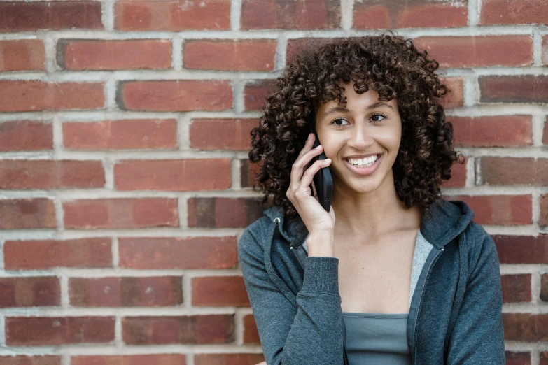 a woman standing in front of a brick wall talking on a cell phone, pexels contest winner, happening, brown curly hair, welcoming smile, varying ethnicities, without text