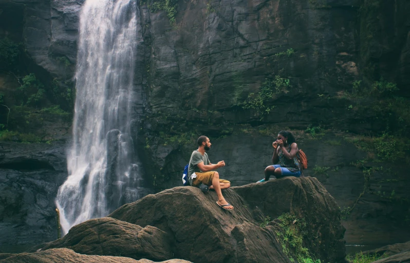 two people sitting on a rock in front of a waterfall, pexels contest winner, people on a picnic, travel guide, sandfalls, thumbnail
