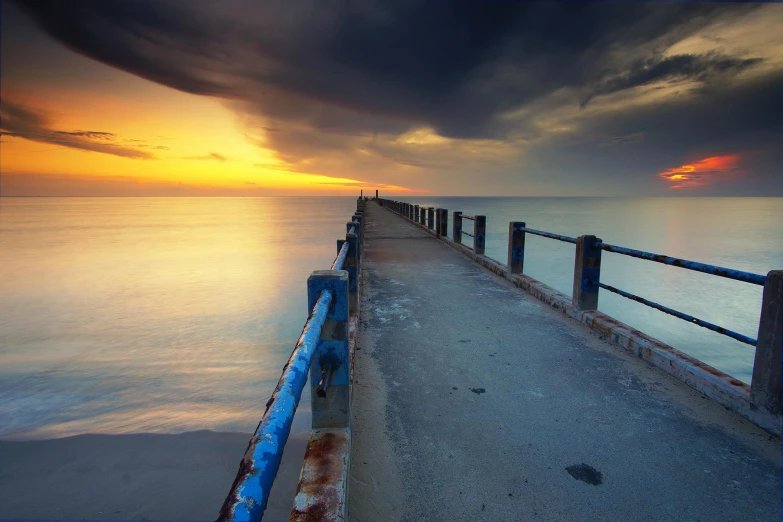 a pier stretching out into the ocean at sunset, a picture, inspired by Jan Rustem, blue and yellow, colour hd photography, mundy, multicoloured