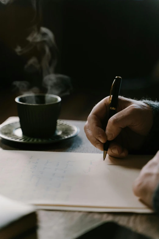 a person writing on a piece of paper next to a cup of coffee, by Sebastian Spreng, paul barson, sydney hanson, cinematic image