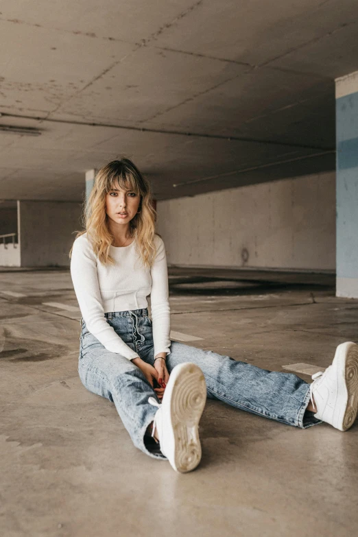 a woman sitting on the ground in a parking garage, an album cover, trending on pexels, sydney sweeney, straight bangs, baggy jeans, in white room