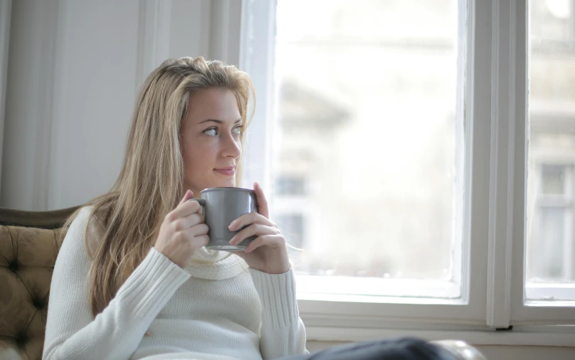 a woman sitting on a couch holding a cup of coffee, by Alice Mason, pexels contest winner, soft grey and blue natural light, looking outside, winter, sitting on a store shelf
