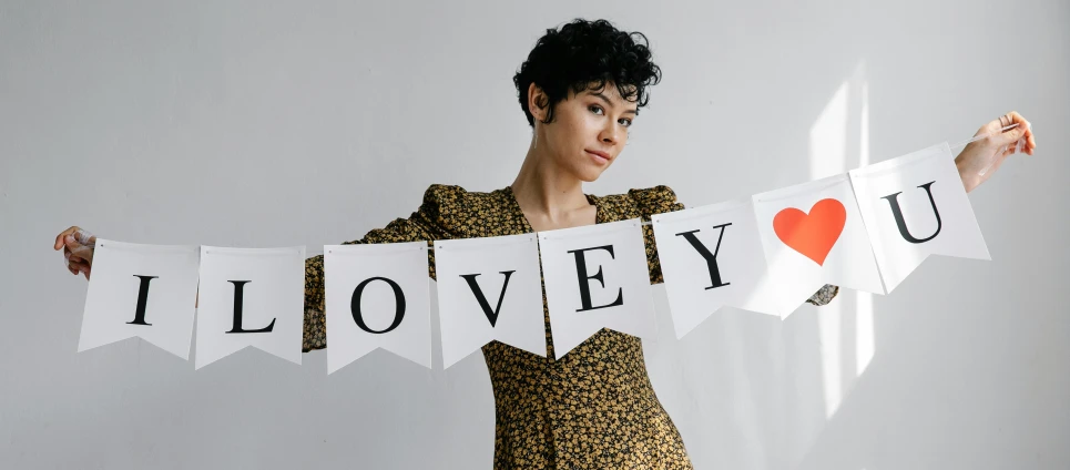 a woman holding a banner that says i love you, an album cover, inspired by Rowena Meeks Abdy, trending on pexels, curly pixie cut hair, fancy clothing, joy ang, \'obey\'