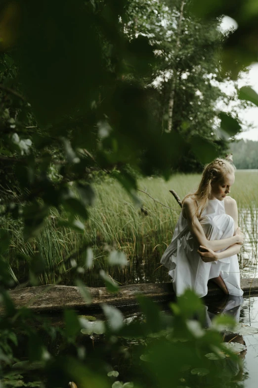a woman sitting on a log in the water, by Grytė Pintukaitė, pexels contest winner, wearing a white folkdrakt dress, maternal, thoughtful, in the wood