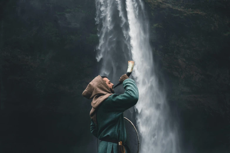 a man standing in front of a waterfall, an album cover, pexels contest winner, hurufiyya, viking attire, drawing an arrow from his quiver, alessio albi, ferred - themed robes and hat