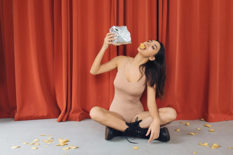 a woman sitting on the floor eating a donut, inspired by Pia Fries, pexels contest winner, magic realism, holly herndon origami statue, wearing a camisole and boots, chips, against a red curtain