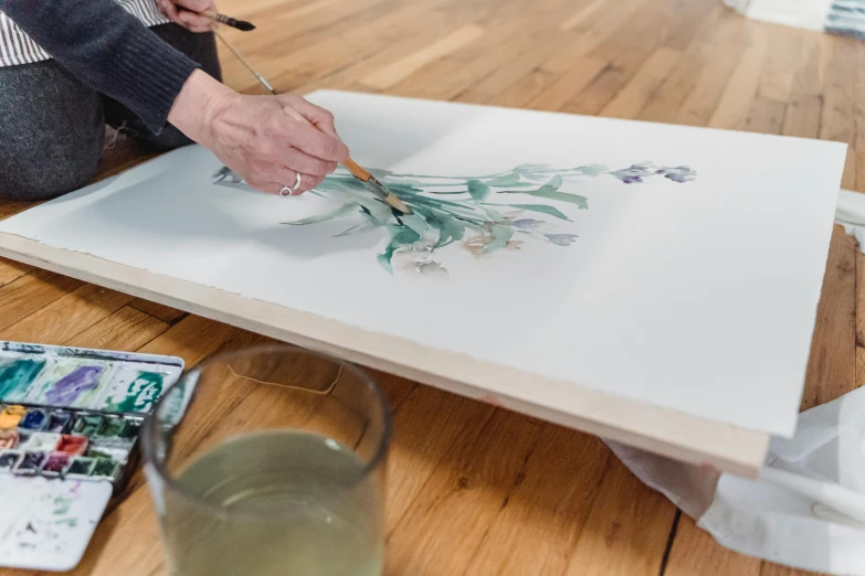 a woman sitting on the floor working on a painting, inspired by Constantin Hansen, featured on unsplash, botanical herbarium paper, on a wooden tray, this painting is a large canvas, on a white table