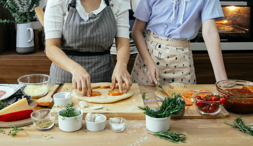 a couple of women standing next to each other in a kitchen, trending on pexels, cooking pizza, herbs, on a wooden table, school class