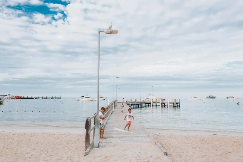 a woman standing on a pier next to a body of water, by Lee Loughridge, pexels contest winner, children playing at the beach, australian beach, light tan, summer street near a beach