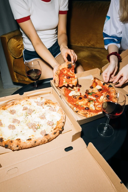 a group of people sitting around a pizza box, on a table, wine, food