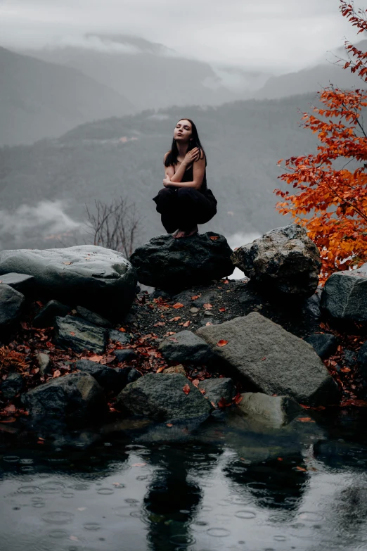 a woman sitting on top of a rock next to a body of water, inspired by Elsa Bleda, pexels contest winner, lovely dark autumn princess, dark grey and orange colours, standing on mountain, thoughtful )