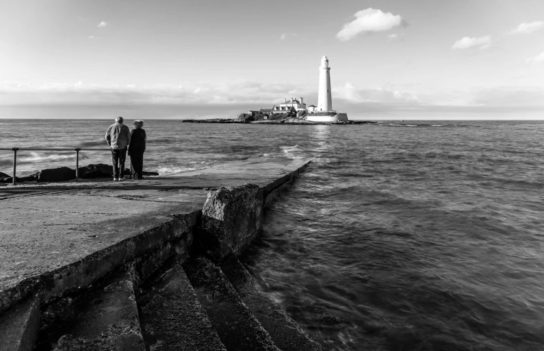 a couple of people standing on top of a pier next to the ocean, a black and white photo, by John Murdoch, pexels, lighthouse, sunken, steps, spire