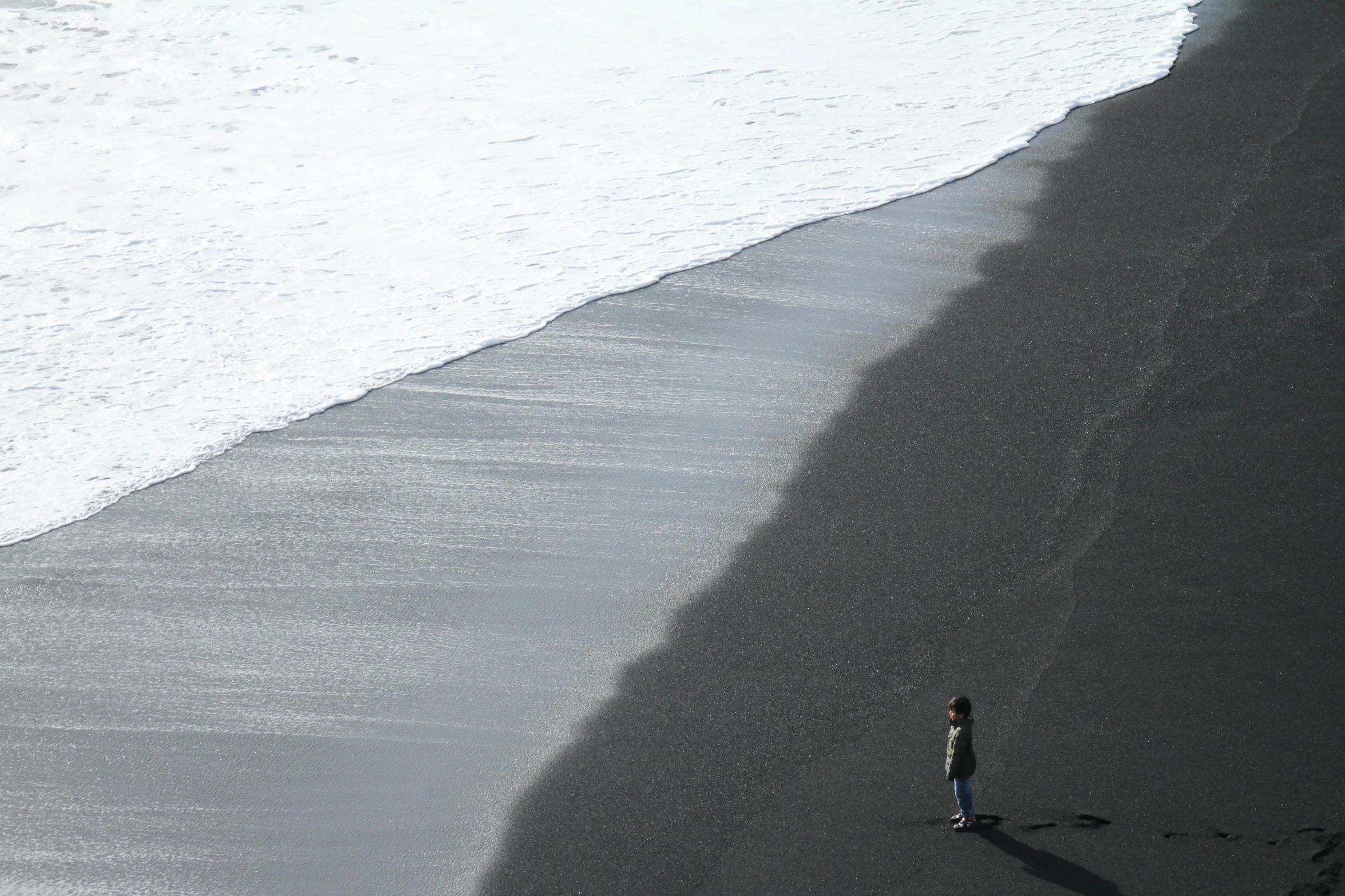 a person standing on a beach next to the ocean, black main color, black sand, from the distance, white beaches