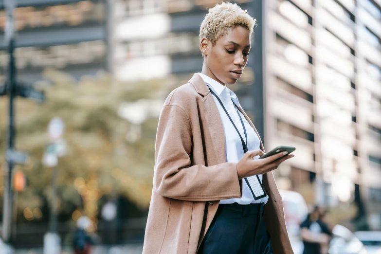 a woman standing on a city street looking at her cell phone, trending on pexels, afrofuturism, bleached blonde short hair, corporate phone app icon, topknot, wearing a blazer