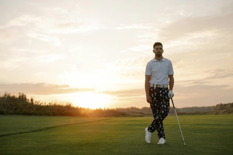 a man standing on top of a green field holding a golf club, a portrait, inspired by Sven Nordqvist, pexels contest winner, patterned clothing, looking off into the sunset, wearing pants and a t-shirt, zayn malik