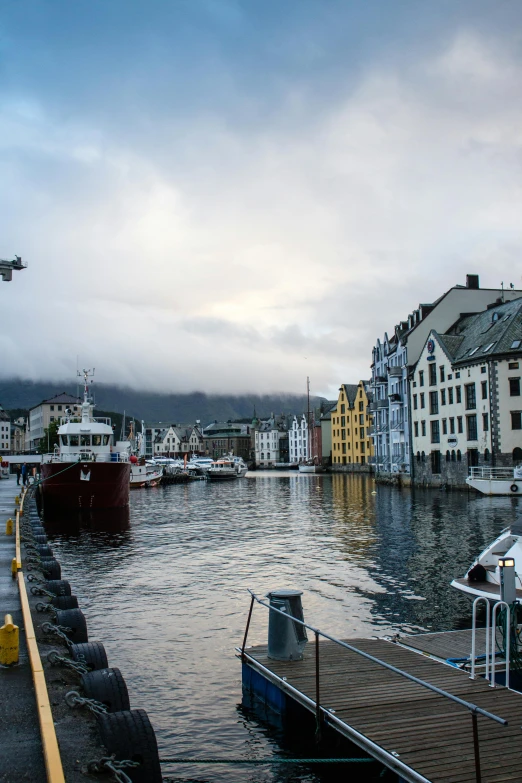 a harbor filled with lots of boats under a cloudy sky, a photo, inspired by Tove Jansson, pexels contest winner, modernism, mountains in the background, square, round buildings in background, canal