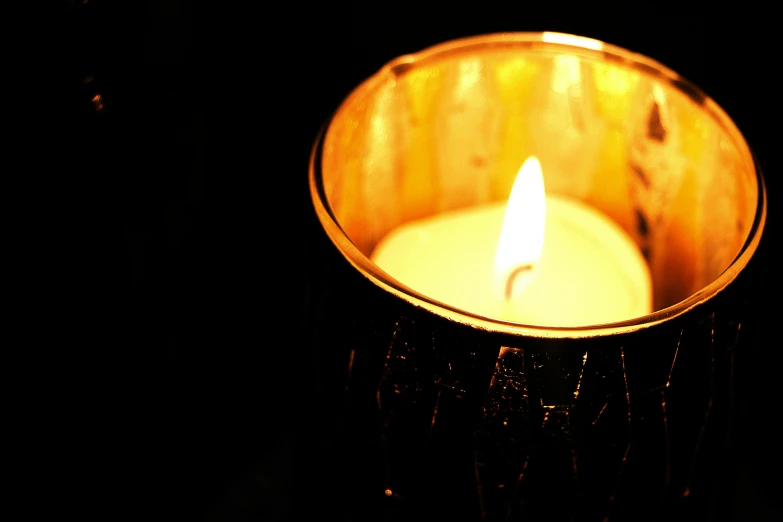 a lit candle sitting on top of a table, glistening gold, dark shading, remembrance, on a candle holder