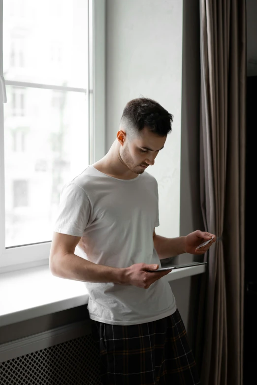 a man standing in front of a window holding a tablet, trending on pexels, renaissance, non binary model, dressed in a white t shirt, concern, in a bedroom