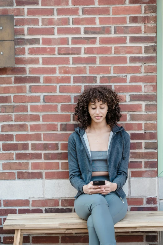 a woman sitting on a bench using a cell phone, by James Morris, trending on pexels, happening, brown curly hair, waiting behind a wall, wearing crop top, gray hoodie