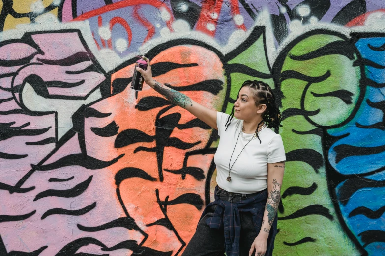a woman standing in front of a graffiti covered wall, by Meredith Dillman, pexels contest winner, graffiti, holding a bottle, hold up smartphone, lorde, people with mohawks