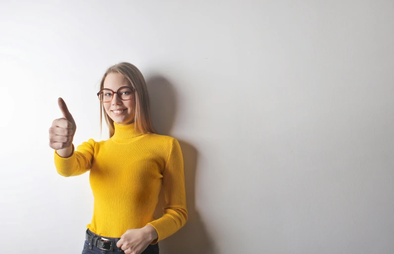 a woman standing in front of a white wall giving a thumbs up, a picture, pexels contest winner, wearing turtleneck, yellow, girl with glasses, holding a candle holder