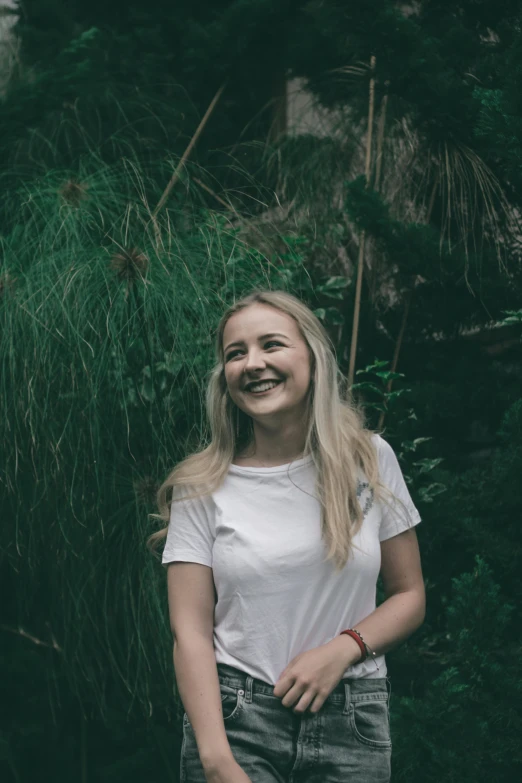 a woman standing in front of a lush green forest, inspired by Elsa Bleda, pexels contest winner, happening, greta thunberg smiling, dressed in a white t shirt, 🤤 girl portrait, young blonde woman