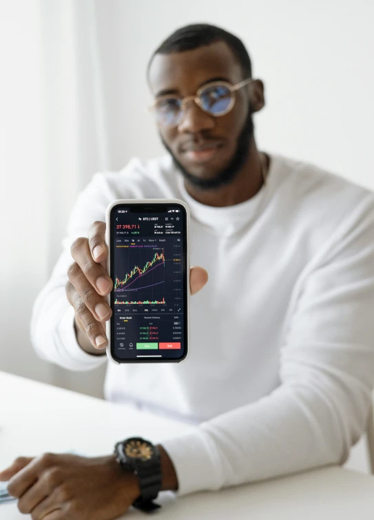 a man sitting at a table holding up a cell phone, displaying stock charts, afro tech, promo image, wētā fx