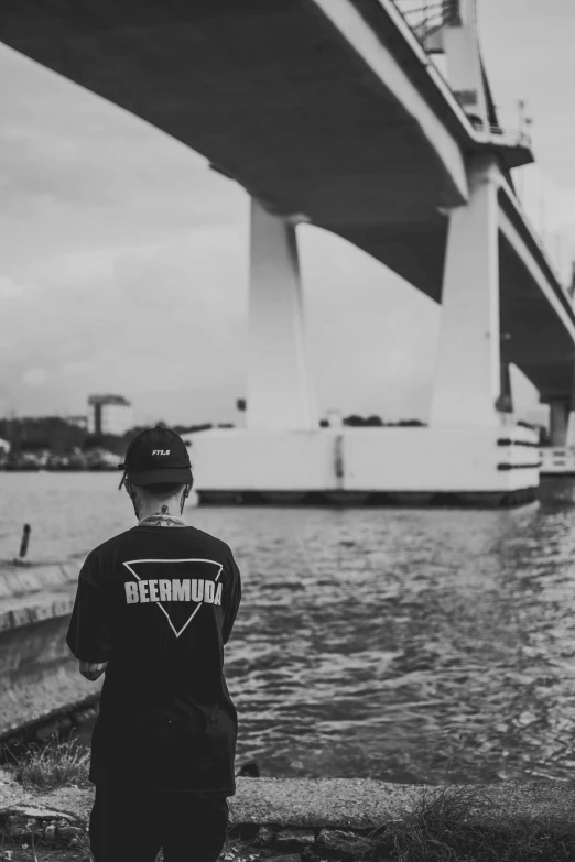 a man standing in front of a body of water, a black and white photo, by Bascove, pewdiepie selfie at a bridge, seen from behind, dreamworld, outlive streetwear collection