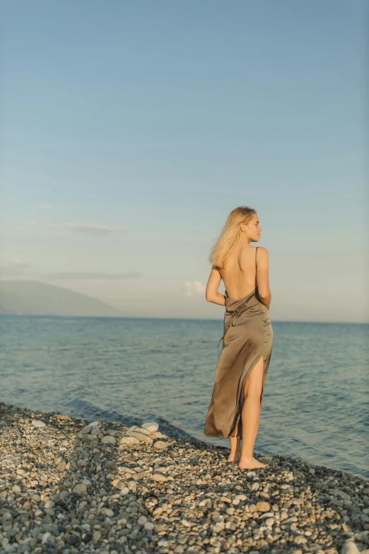 a woman standing on a rocky beach next to the ocean, open back dress, aphrodite, a blond, sun down