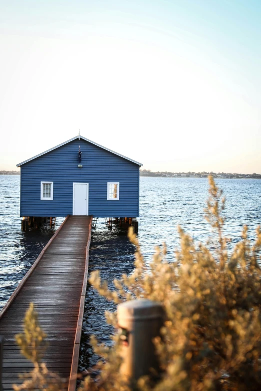 a blue house sitting on top of a body of water, by Peter Churcher, hut, at golden hour, william warehouse, body of water