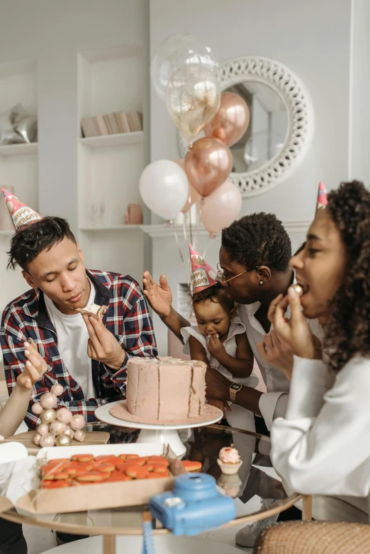 a group of people sitting around a table with a cake, balloons, on a white table