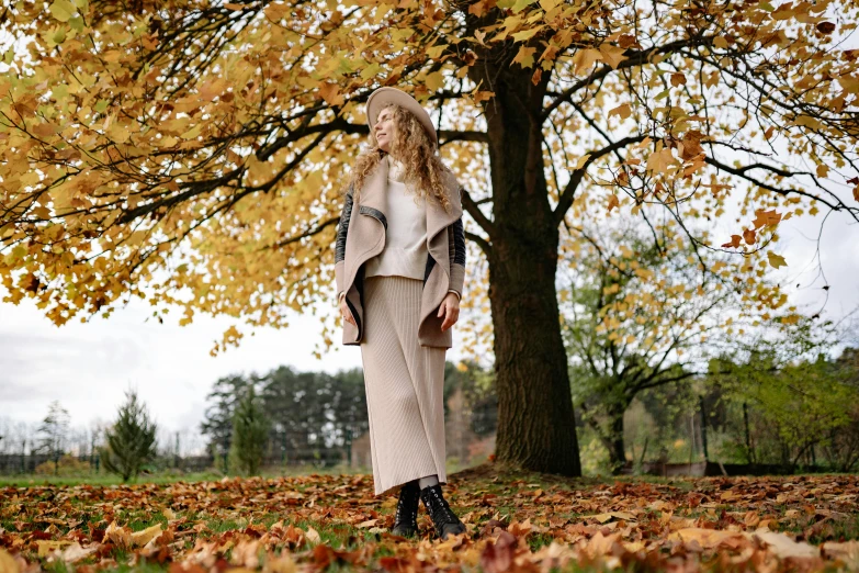 a woman standing under a tree in a park, by Emma Andijewska, pexels, light brown coat, layered skirts, thumbnail, fall