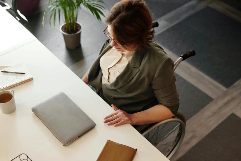 a woman sitting at a table working on a laptop, trending on pexels, sitting in a wheelchair, top-down shot, female in office dress, thumbnail