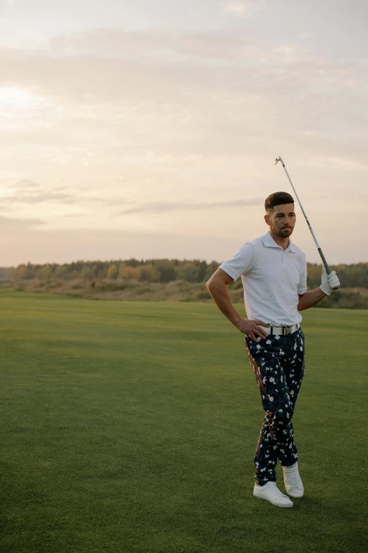 a man standing on top of a green field holding a golf club, a portrait, inspired by Sven Nordqvist, pexels contest winner, trout in pants, patterned clothing, evening time, dasha taran