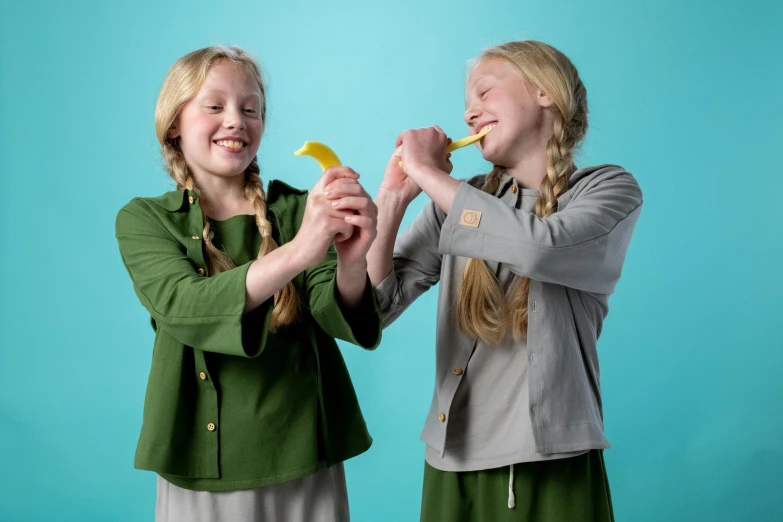 two young girls brushing their teeth against a blue background, inspired by Elsa Beskow, hurufiyya, giant bananas natural disaster, wearing green jacket, official product photo, grey