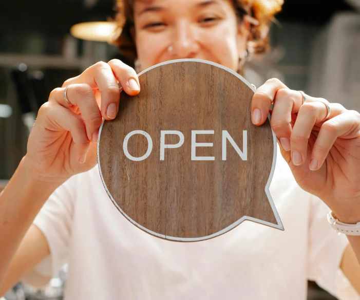 a woman holding an open sign in front of her face, trending on pexels, award winning shopfront design, on a wooden tray, portal opening, brown