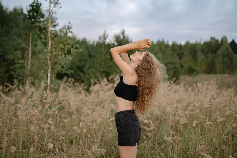 a woman standing in a field of tall grass, pexels contest winner, croptop and shorts, frizzy hair, dance meditation, near forest