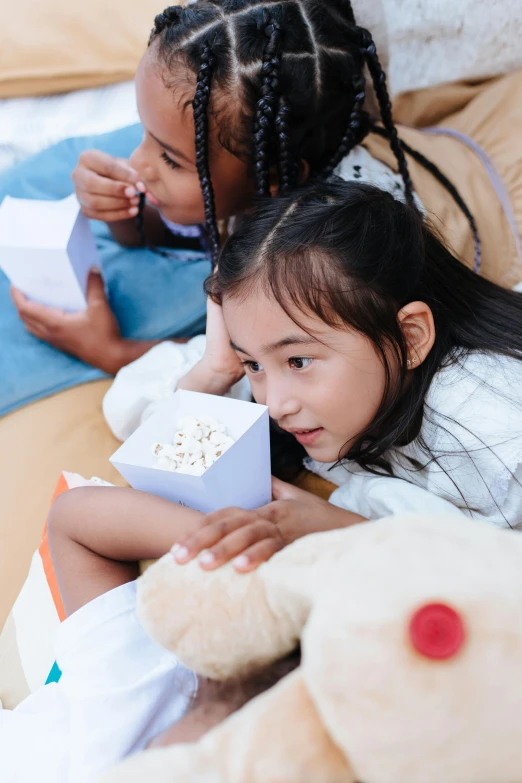 a couple of little girls laying on top of a bed, a cartoon, pexels contest winner, sitting in a movie theater, young asian girl, having a snack, square