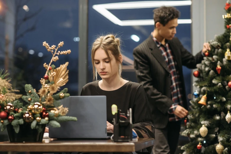 a woman sitting in front of a laptop computer next to a christmas tree, by Emma Andijewska, pexels contest winner, renaissance, in a open-space working space, medium shot of two characters, standing elegantly, busy night