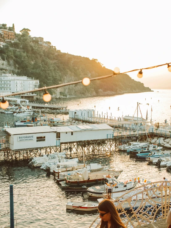 a woman sitting in a hammock next to a body of water, by Julia Pishtar, pexels contest winner, renaissance, harbour in background, capri coast, warm glow from the lights, wood pier and houses