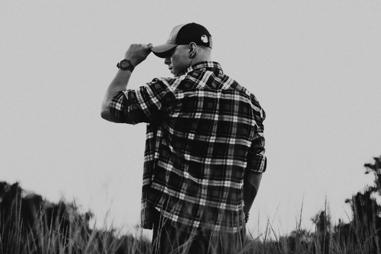 a man standing in a field of tall grass, a black and white photo, by Austin English, pexels, lumberjack flannel, john cena, profile pic, wearing a backwards baseball cap