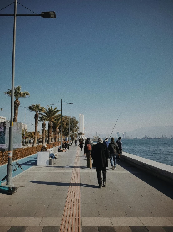 a group of people walking down a sidewalk next to a body of water, the sea seen behind the city, background image, fishing, aykut aydogdu